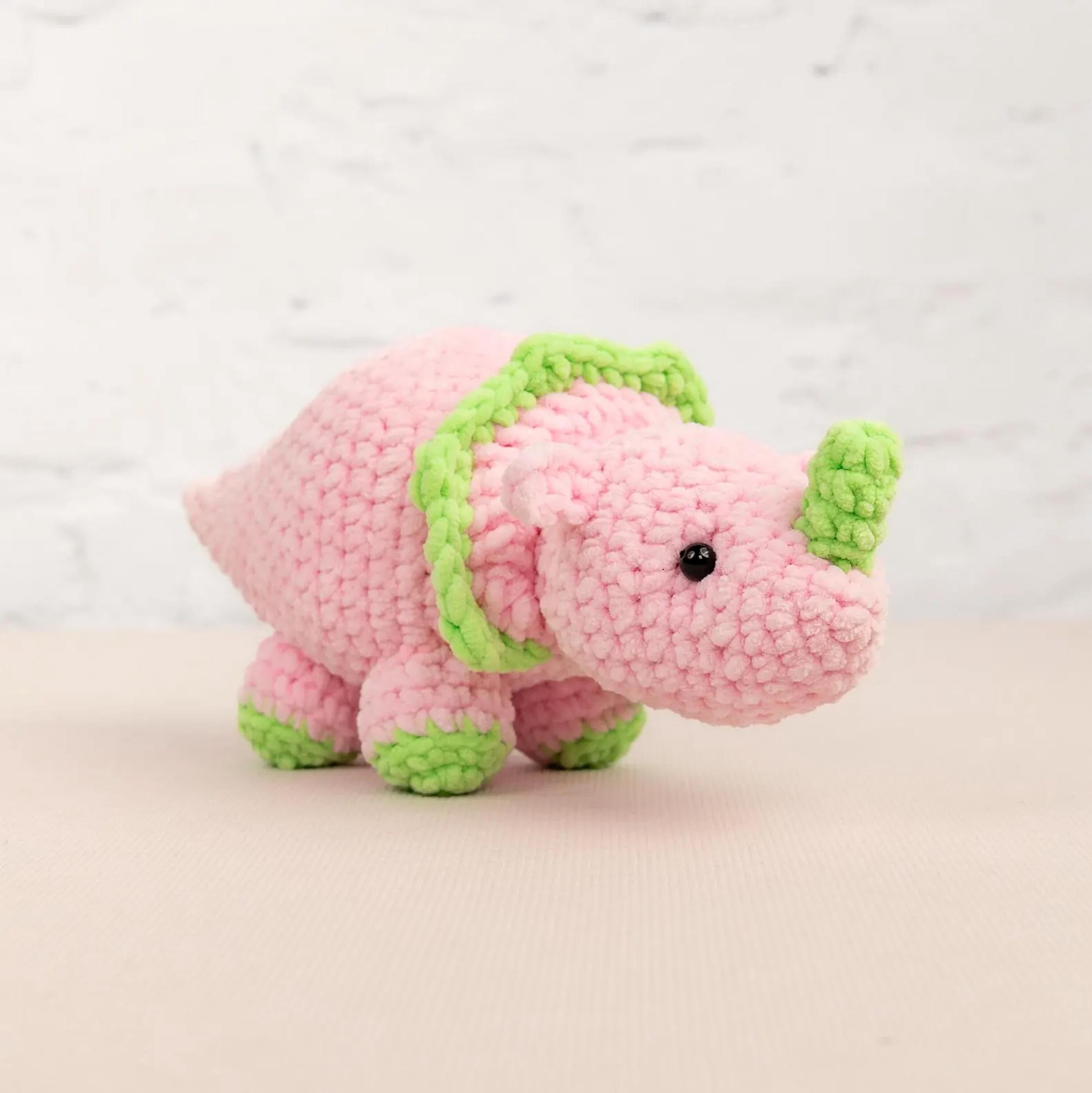 A pink crocheted toy dinosaur with green accents, sitting on a light-colored surface against a white background.